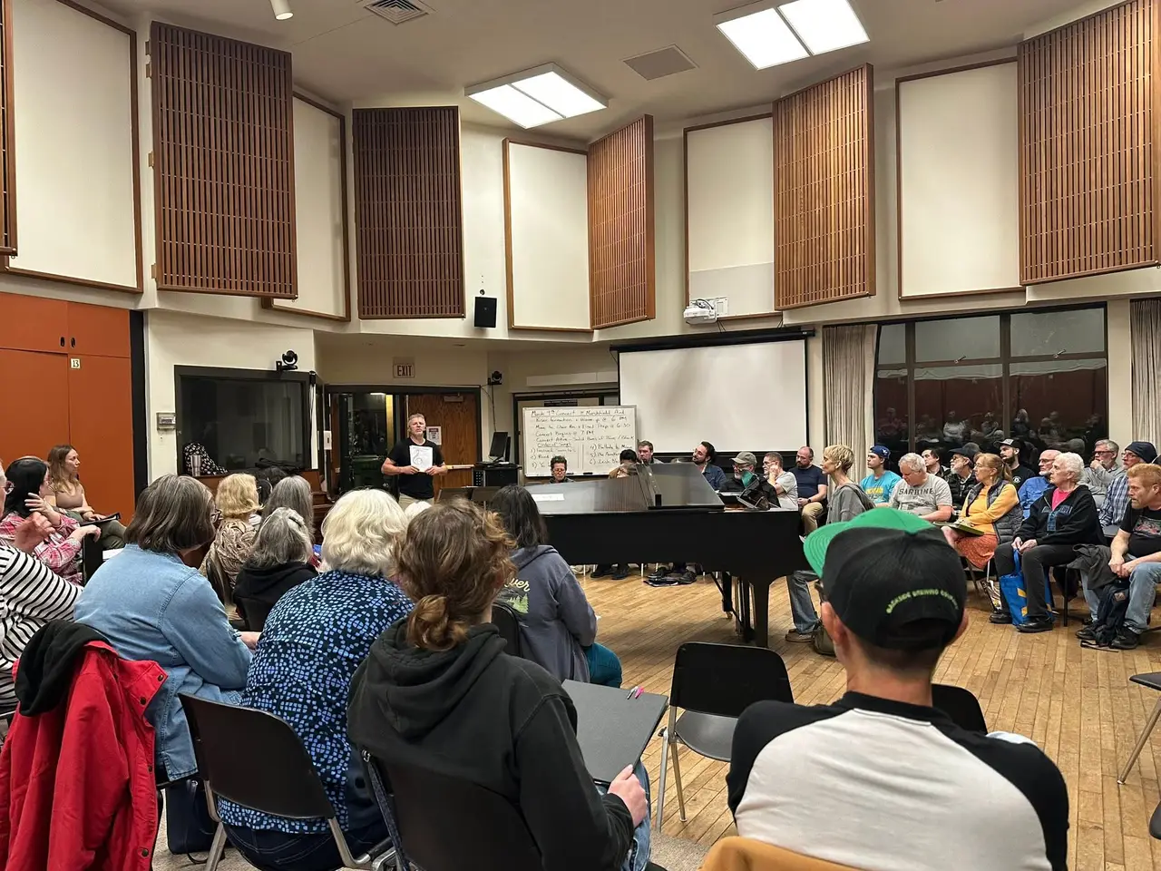 The full choir rehearses in a circle around a grand piano, concert plan on the whiteboard.