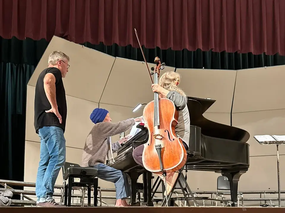 A cellist and pianist rehearse at a grand piano while the director looks on.